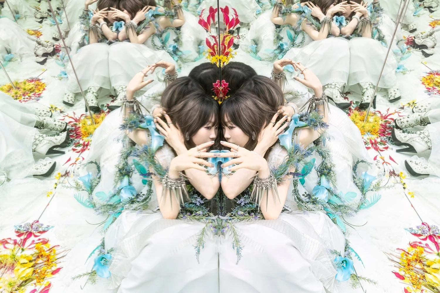 白いドレスをまとった女性が万華鏡のような反射効果で描かれた幻想的な画像です。青い花や緑の植物、カラフルな花が散りばめられ、シュールで美しい世界観を表現しています。中央では二人の女性が向かい合っています。