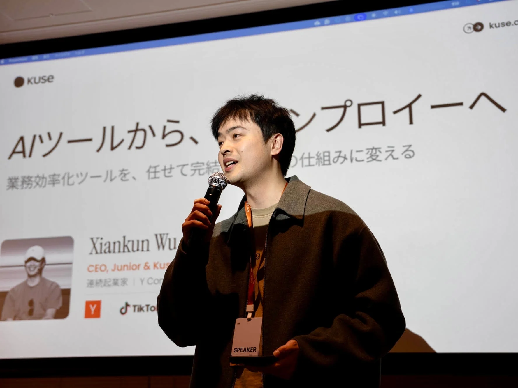 「AIツールから、エンプロイーへ」と書かれたスクリーンを背景に、マイクを持って講演する男性