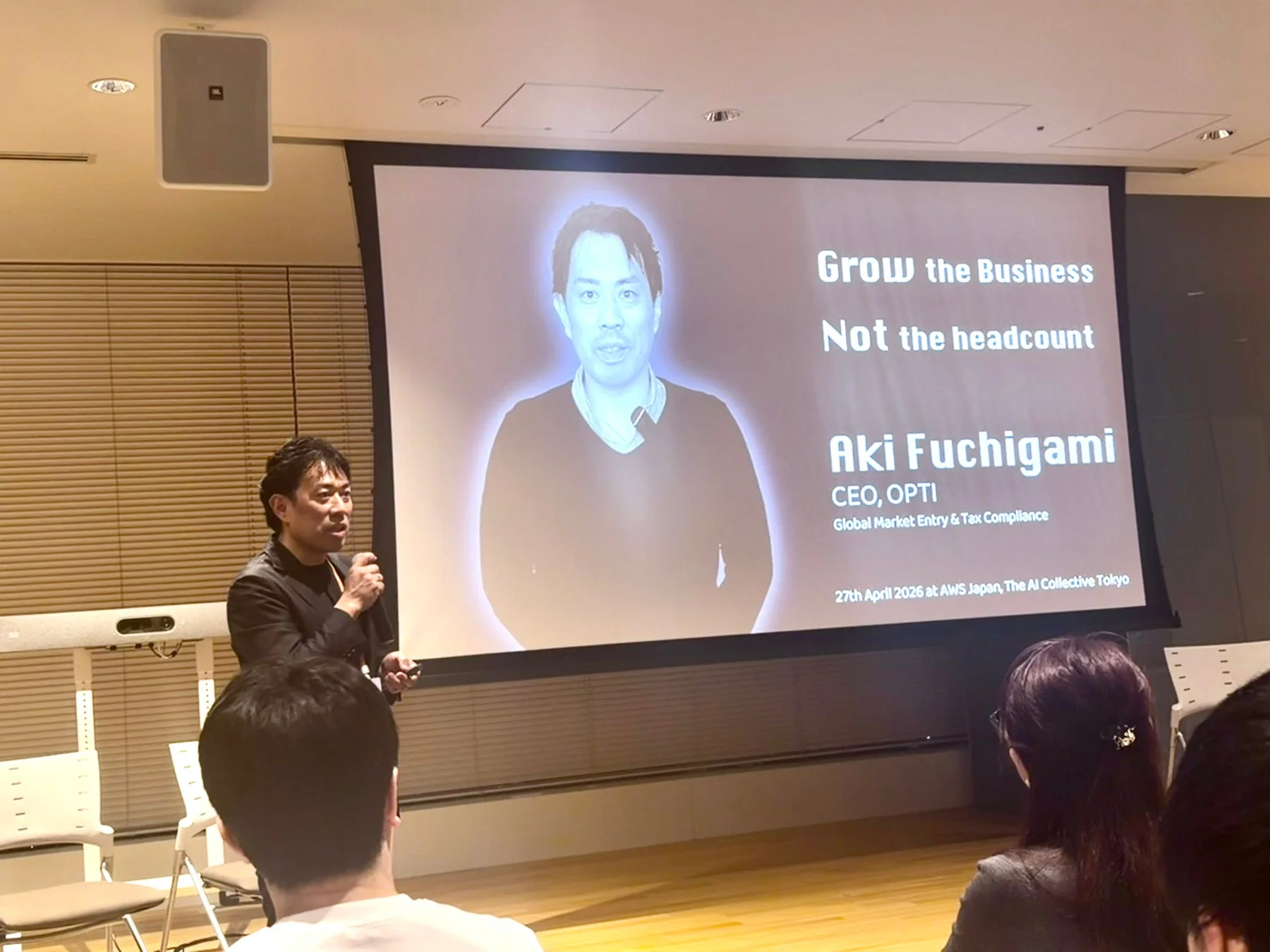 ビジネス成長に関する講演会で、講師がスクリーンに映し出された自身の情報と「Grow the Business Not the headcount」というスローガンを背景に話している