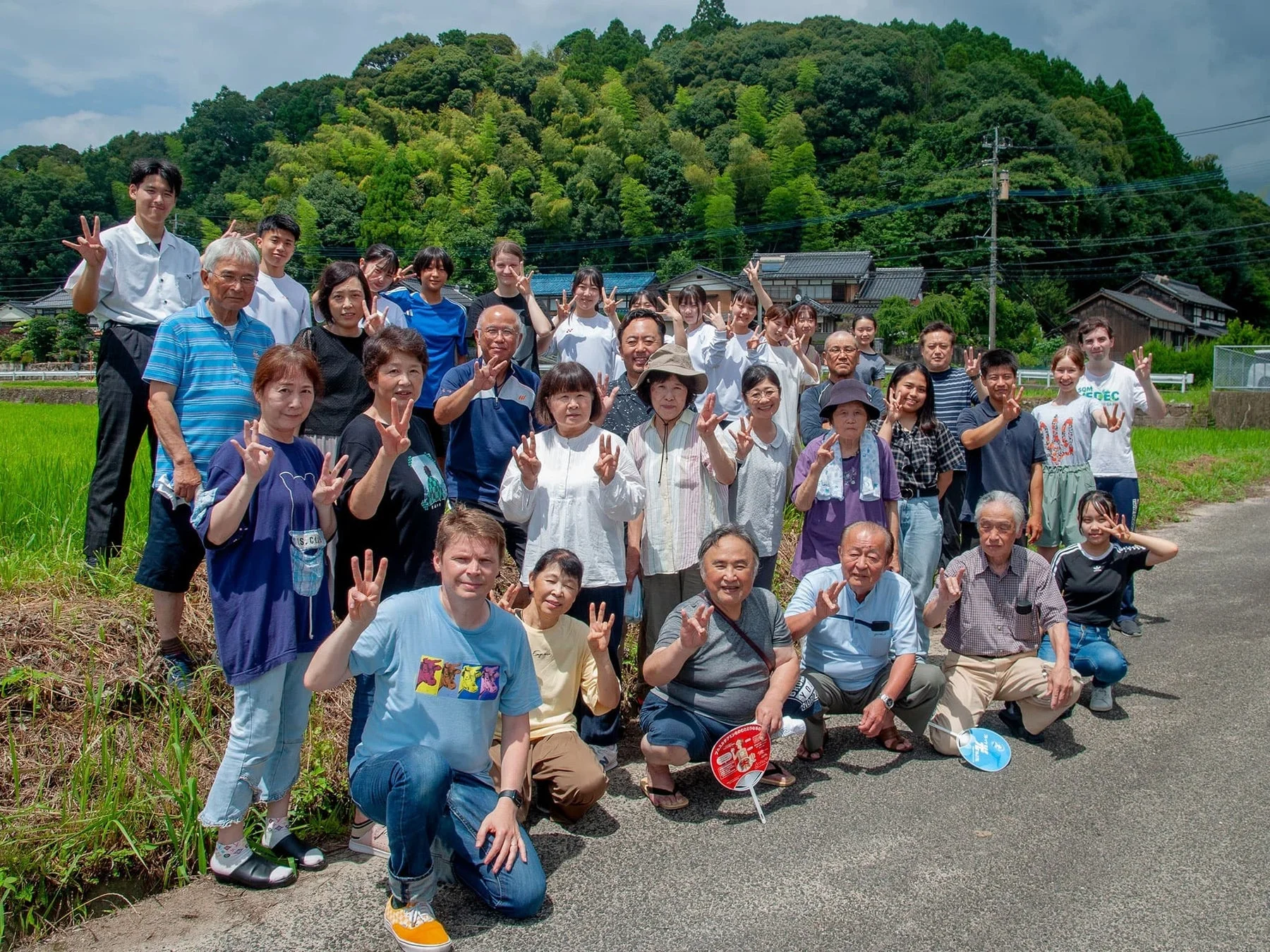 多世代の地域住民と外国人が集まり笑顔でピースサインをする集合写真