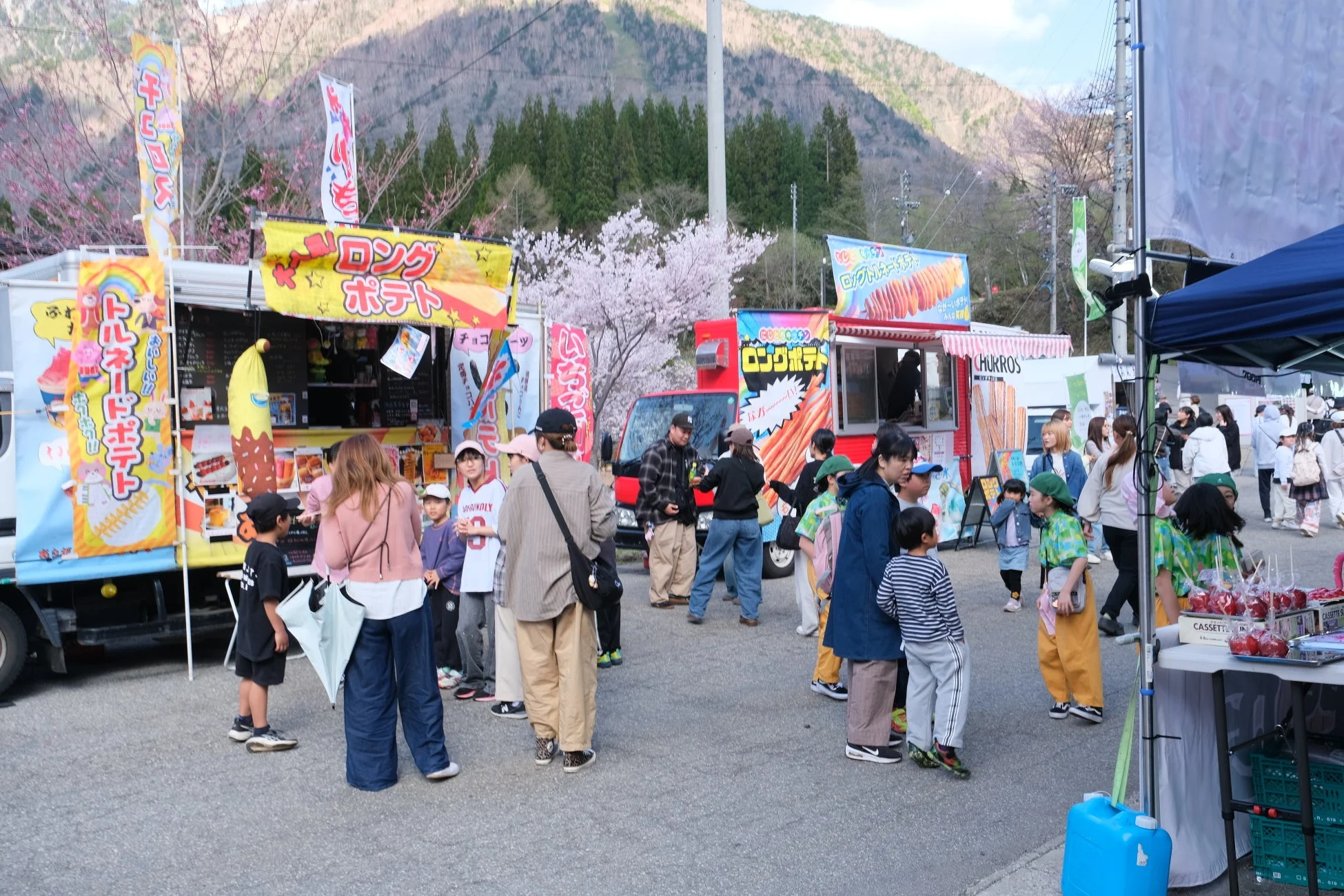 山を背景に桜が咲く屋外イベント会場で、多くの人々、特に家族連れや子供たちが集まっています。ロングポテトやトルネードポテト、チュロスなどを販売するキッチンカーが並び、賑わいを見せています。