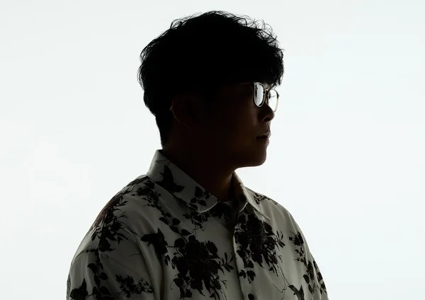 白い背景を背に、メガネをかけた男性の横顔がシルエットで写っている。柄物のシャツを着用し、クールで神秘的な雰囲気を醸し出しているポートレート写真。