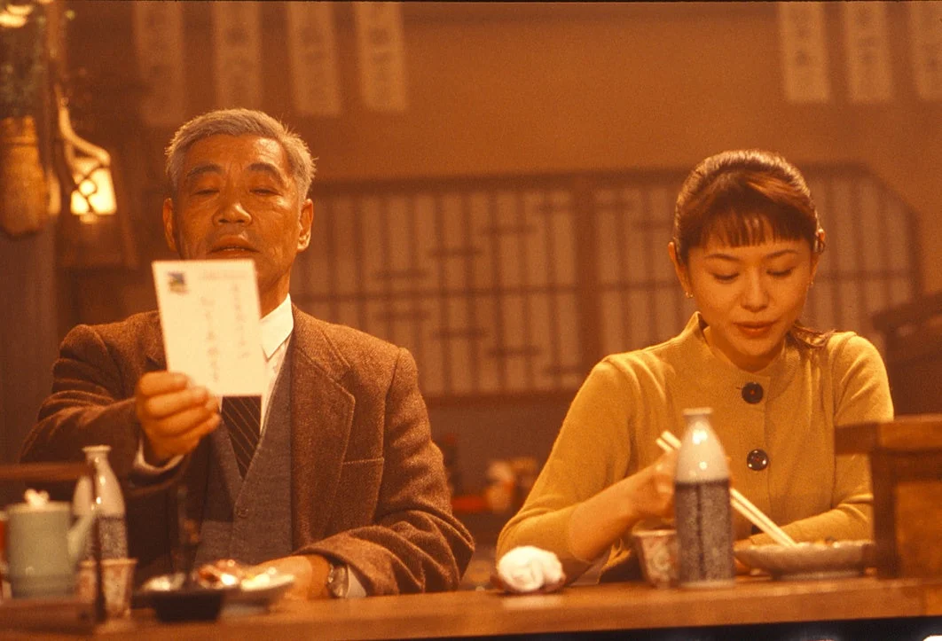 温かい雰囲気の店内で、年配の男性が何か書類を見せながら、食事中の若い女性とカウンター越しに話している