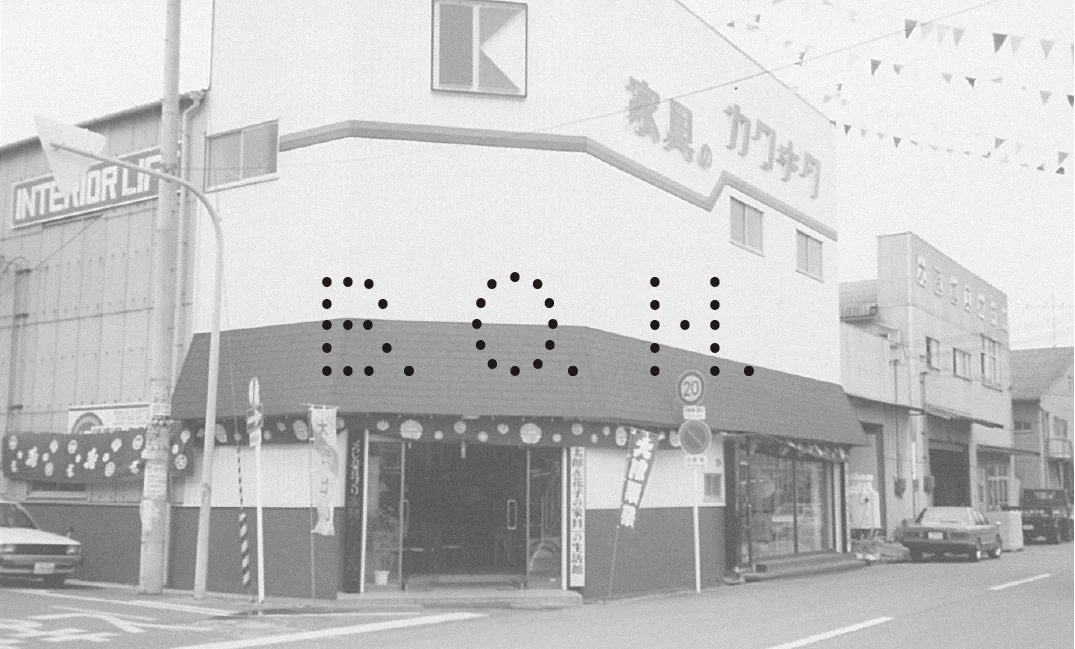 昭和レトロな雰囲気の日本の街角にある店舗の白黒写真です。角地の建物には「家具」と「INTERIOR LI」の看板が見え、通りには車が停まっています。上空には旗飾りがあり、日常の風景が写し出されています。
