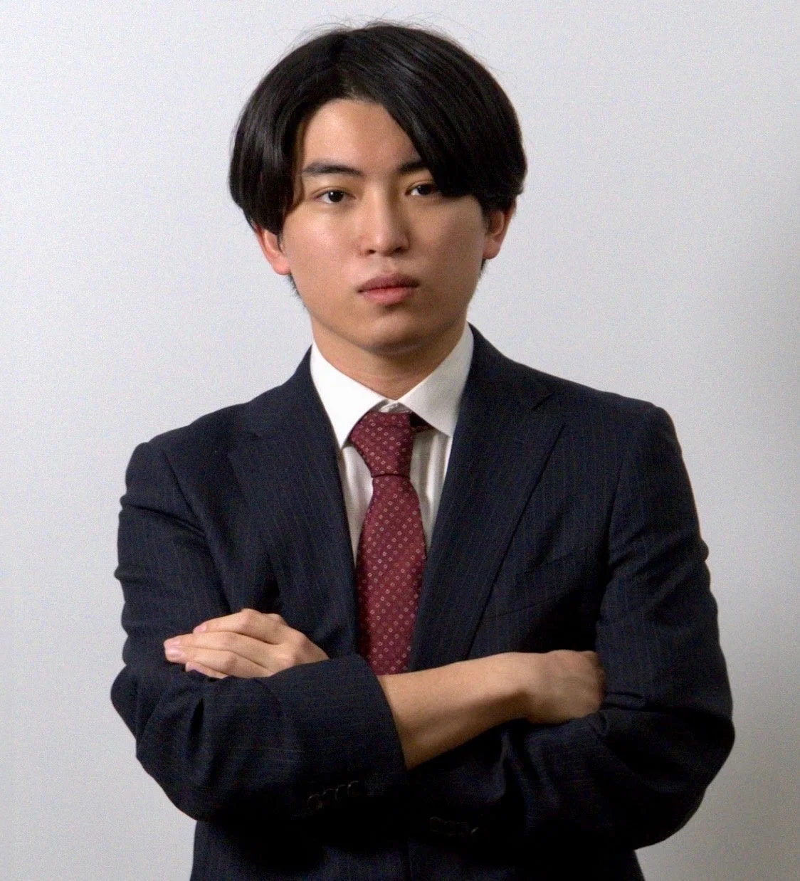 白い背景の前で腕組みをして立つ、スーツとネクタイを着用した若い男性のポートレート写真です。真剣な表情でカメラを見つめており、ビジネスシーンでの利用に適しています。