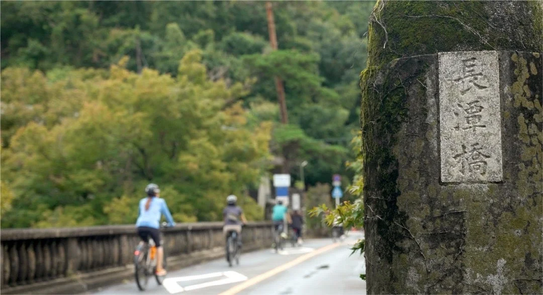 苔むした柱に「長澤橋」と刻まれた石板があり、数人のサイクリストが橋の上を自転車で走る風景