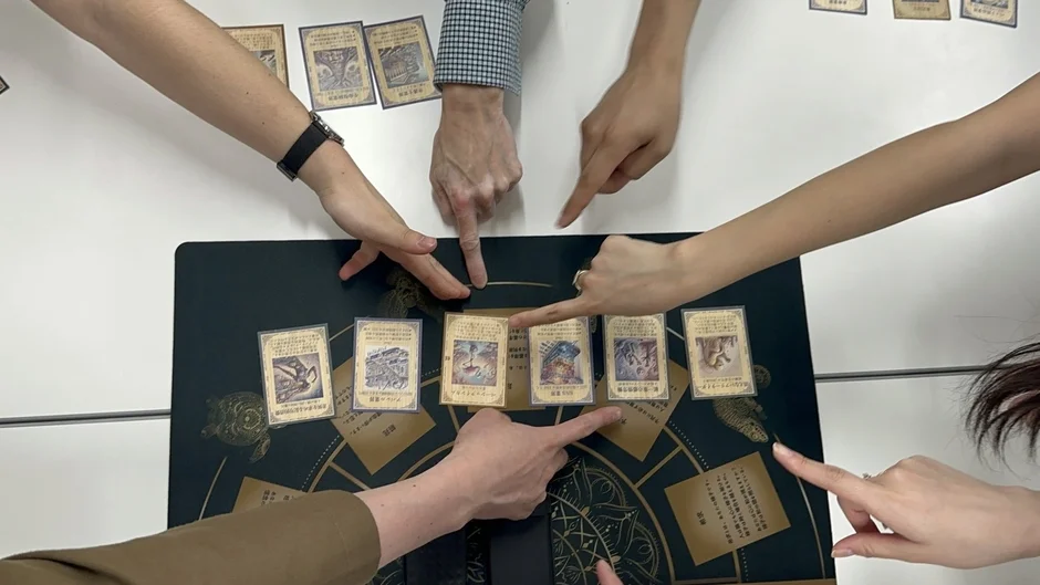 複数の人がテーブルを囲み、ダークグリーンのプレイマットの上に並べられたカードを指差しながらカードゲームを楽しんでいる様子