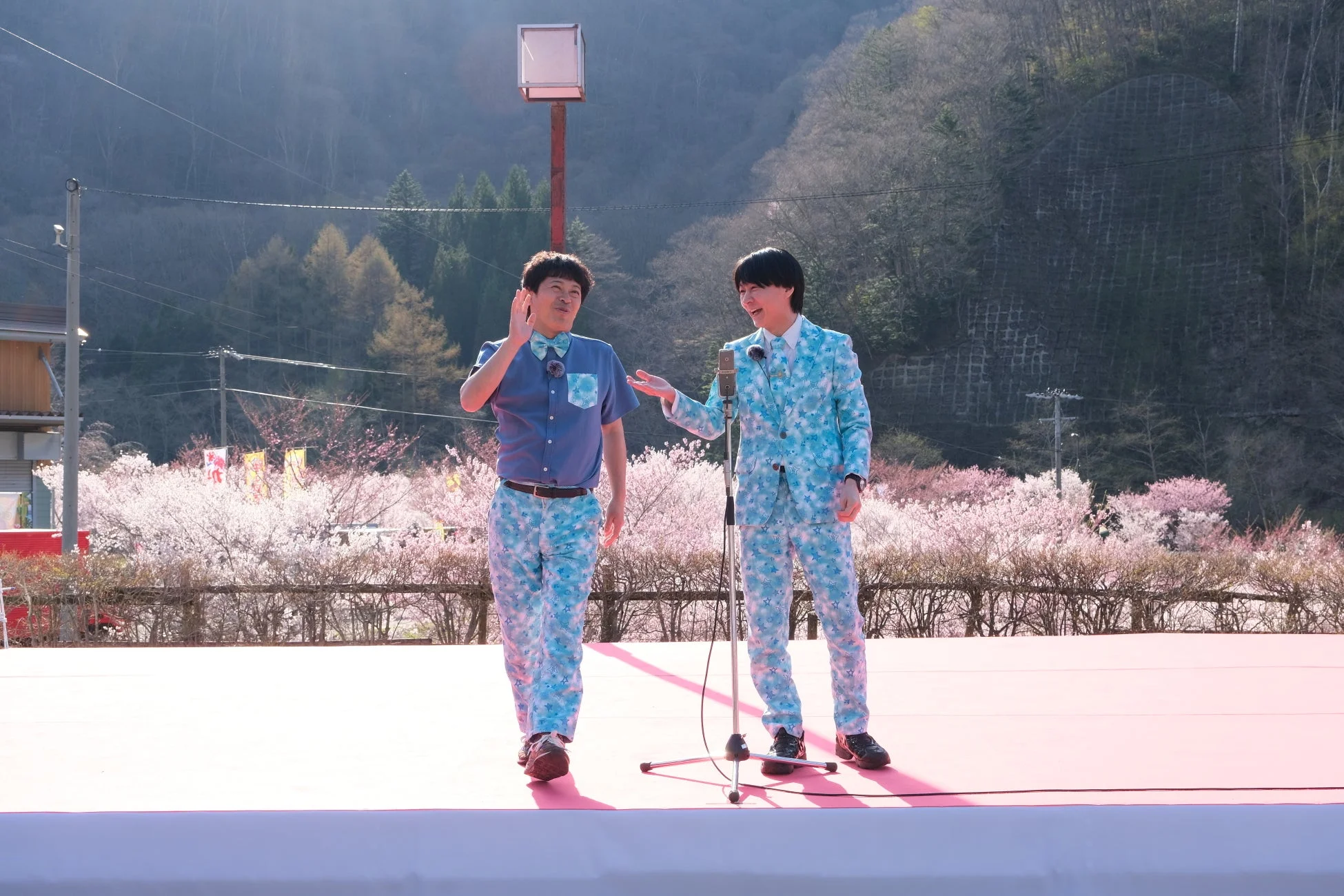 春の桜が満開の屋外ステージで、星柄の衣装を着た男性二人組がマイクを持ってパフォーマンスを行っています。背景には山と桜並木が広がり、イベントの賑やかな雰囲気が伝わってきます。
