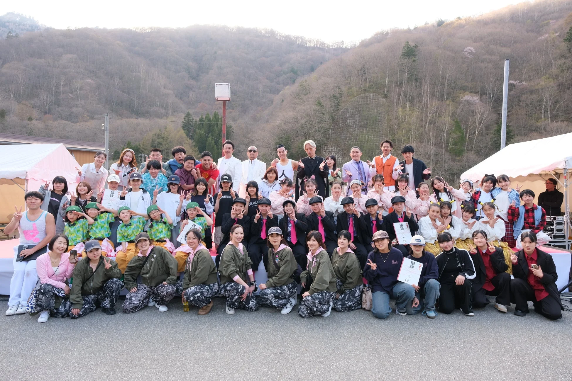 屋外のイベント会場で、様々な衣装を着た大人と子供が大勢で集合写真を撮っている様子。多くの人が笑顔でポーズをとり、中には賞状を持つ人もいる。山を背景に、楽しそうな雰囲気が伝わる一枚。