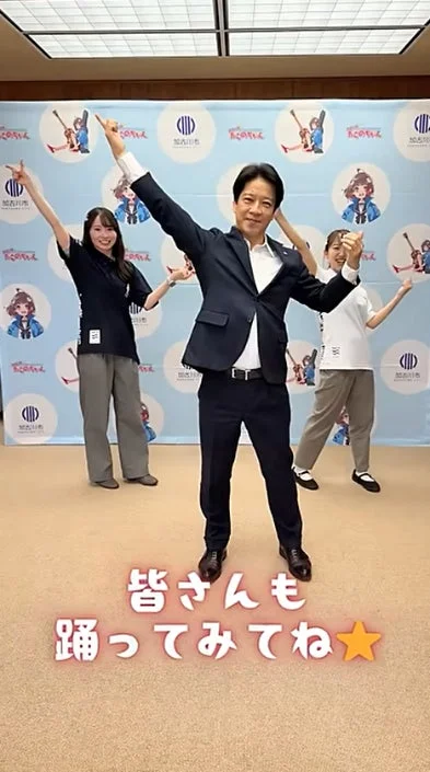 スーツ姿の男性と2人の女性が加古川市のキャラクターの前で踊る様子
