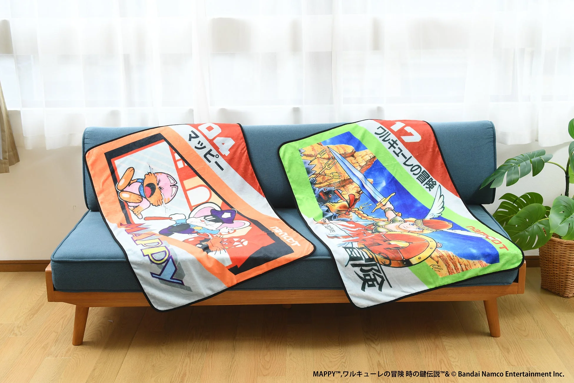 青いソファに、レトロゲーム「マッピー」と「ワルキューレの冒険」のキャラクターがデザインされたブランケットが2枚かけられています。リビングの窓際で、観葉植物も置かれた落ち着いた雰囲気です。