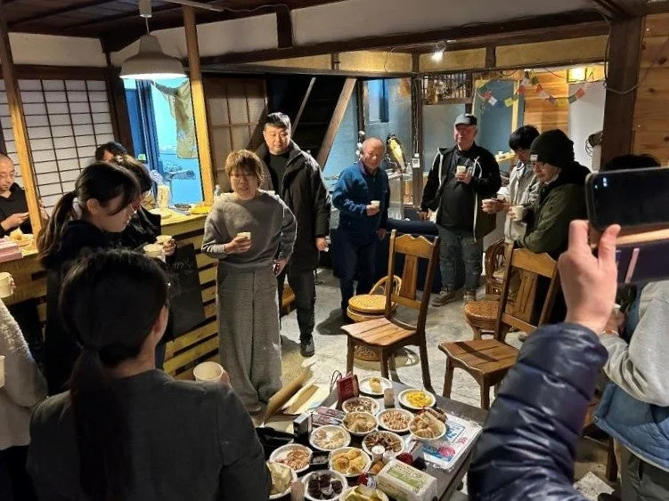 古民家風の屋内で、多くの人々が集まって歓談している様子。中央のテーブルには様々な料理や飲み物が並べられており、カジュアルな交流会やパーティーが開催されているようだ。