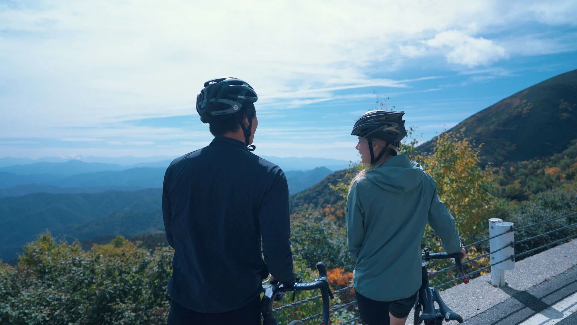 山頂付近の道路で自転車に乗り、遠くの山々を見渡す男女のサイクリスト
