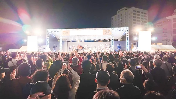 夜の屋外ステージで行われているライブイベントで、多くの観客がステージに向かって拍手を送ったり、スマートフォンで撮影したりしている様子。ステージは明るくライトアップされ、熱気あふれる雰囲気が伝わってきます。