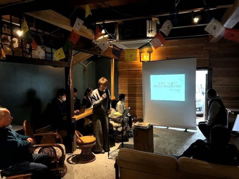 「自然ビール(仮称)の研究会」と題されたプレゼンテーションが行われている室内のイベント風景。登壇者がマイクを使い、参加者に向けて説明している。ルンタが飾られた独特の空間で、多くの人が熱心に耳を傾けている。