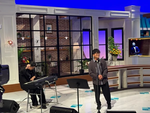 音楽スタジオのセットで、一人の男性がマイクの前で歌い、もう一人の男性がキーボードを演奏している。テレビ番組のリハーサルか収録風景のようだ。