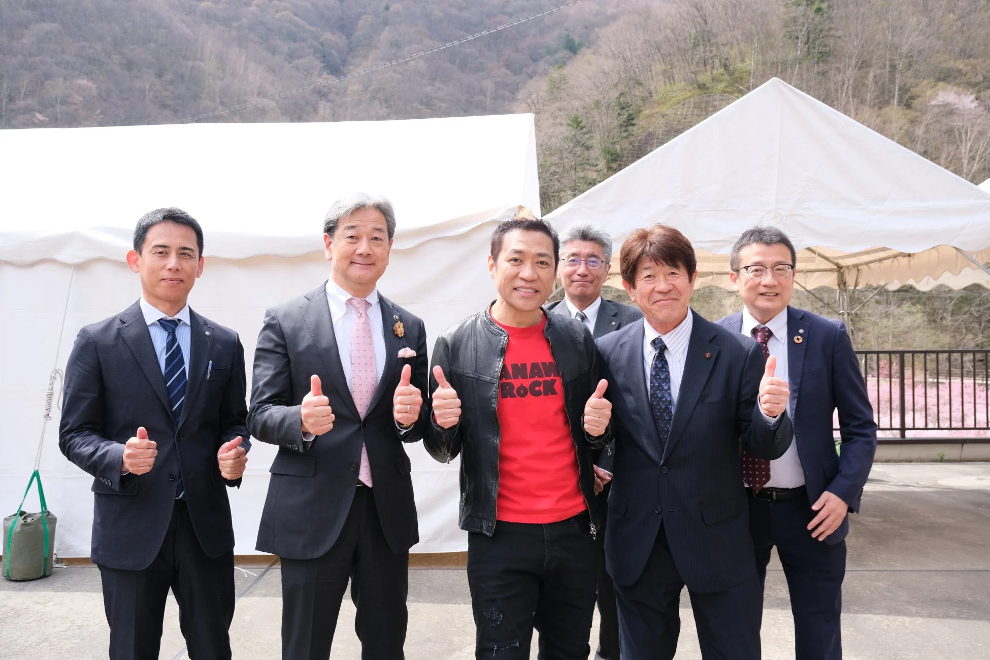 屋外で6人の男性が笑顔でサムズアップしている集合写真です。ほとんどの男性はスーツを着用しており、背景には山や白いテント、ピンクの花が見えます。和やかな雰囲気のイベント風景です。