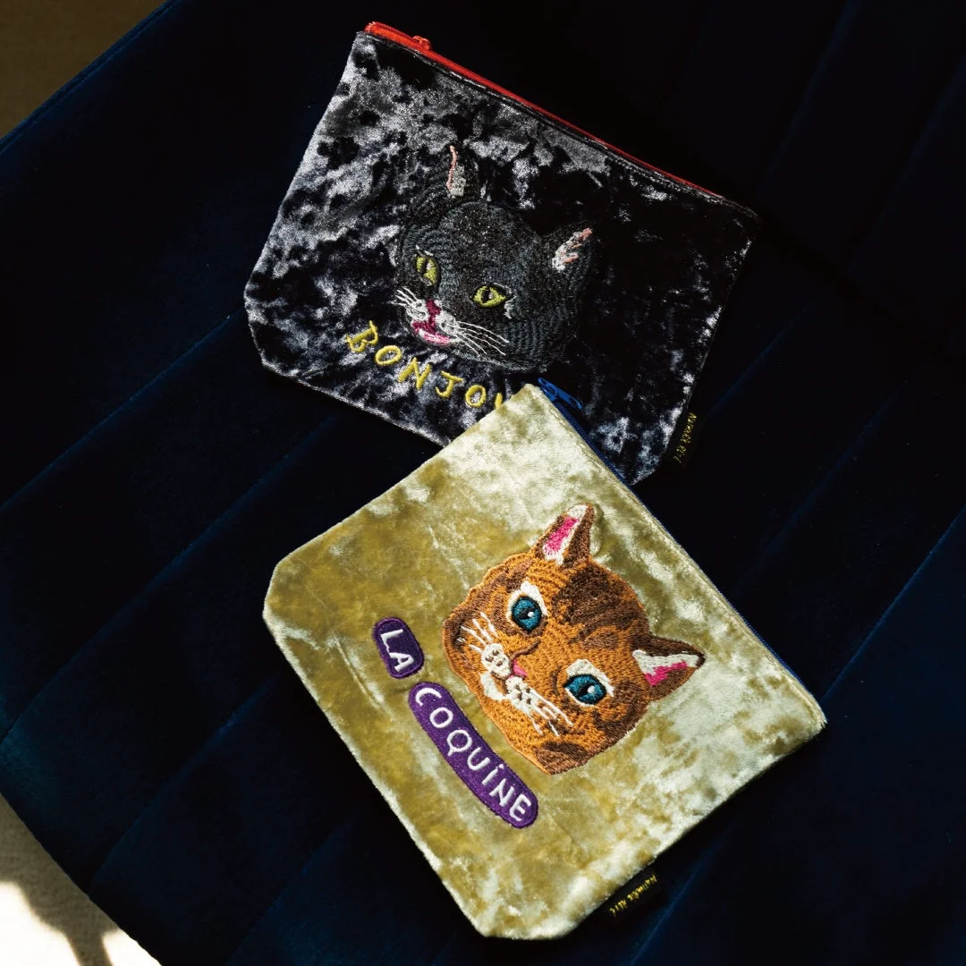 黒いベルベット生地に黒猫の顔と「BONJOUR」の刺繍が施されたポーチと、黄緑色のベルベット生地に茶トラ猫の顔と「LA COQUINE」の刺繍が施されたポーチが、暗い青色の背景に置かれています。猫好きにはたまらない可愛らしいデザインのファッション小物です。