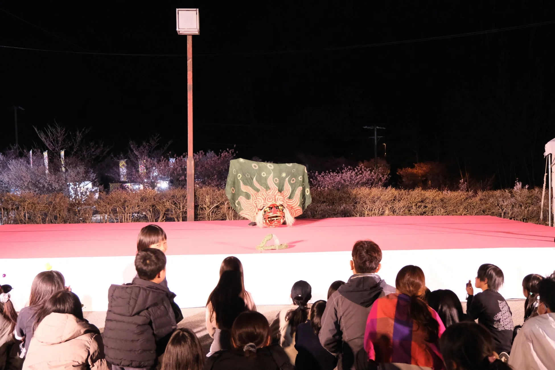 夜間の屋外ステージで、多くの観客が伝統的な獅子舞のパフォーマンスを鑑賞している様子です。背景にはライトアップされた木々が見えます。