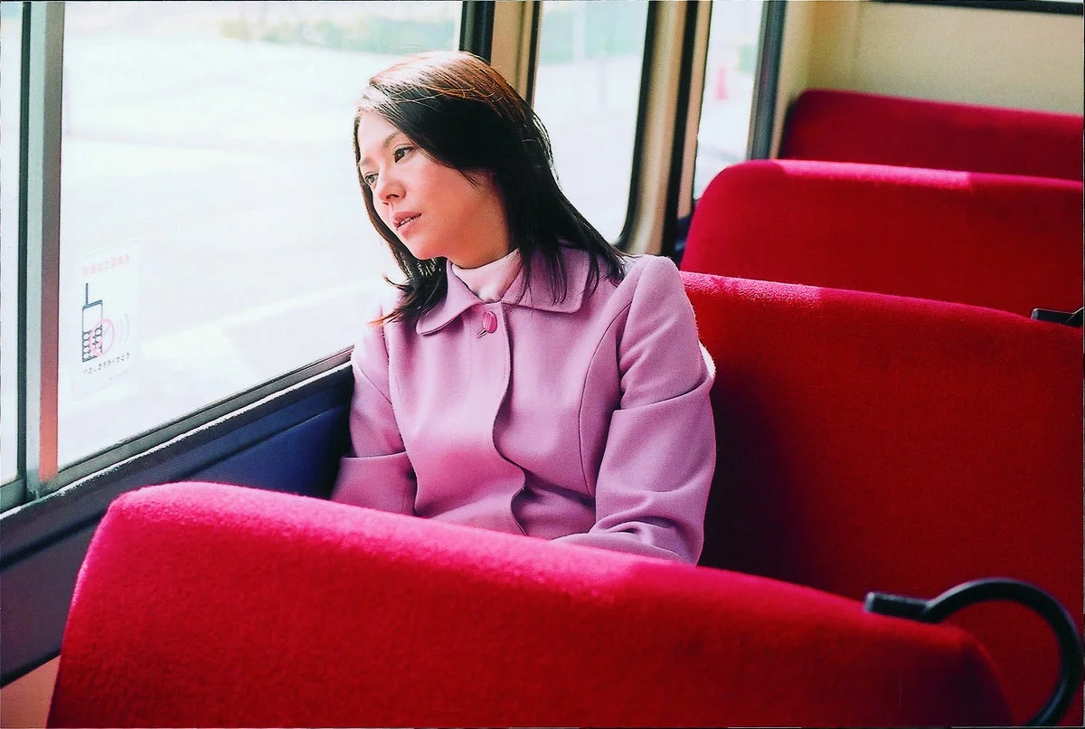 ピンクのジャケットを着た女性が電車かバスの赤い座席に座り、窓の外をぼんやりと眺めている様子