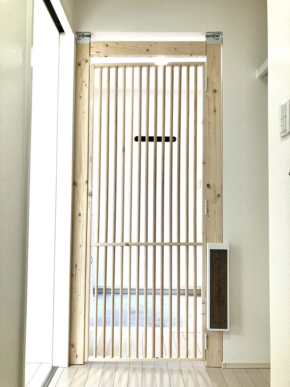 部屋の入り口に設置された、明るい木製の縦格子状のドアです。ペットゲートとしても機能し、隣には猫用の爪とぎも備え付けられています。