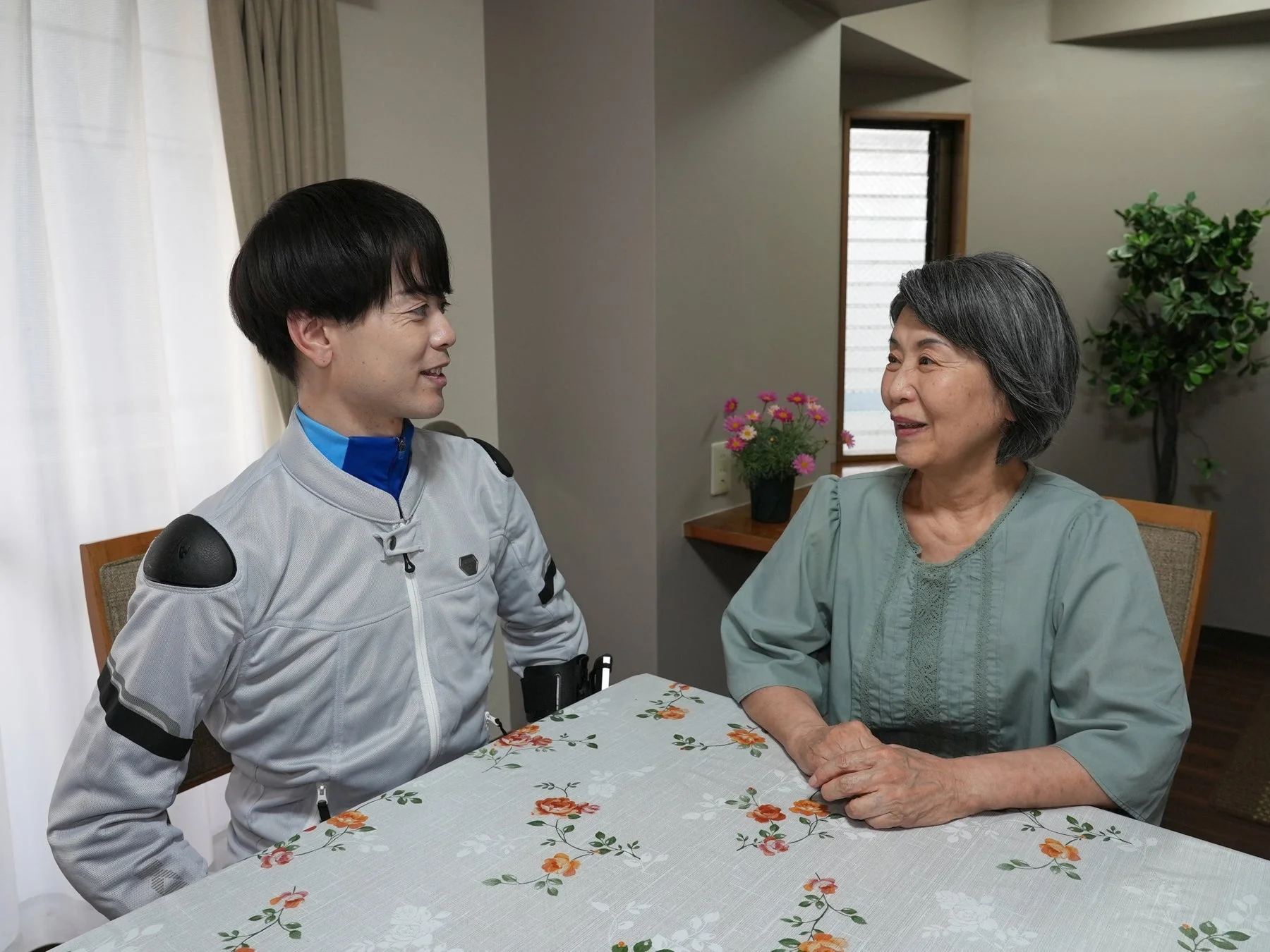 若い男性と高齢の女性が、花柄のテーブルクロスが敷かれたテーブルを挟んで向かい合い、笑顔で会話している様子です。和やかな雰囲気の室内で、二人の間に温かい交流が見られます。