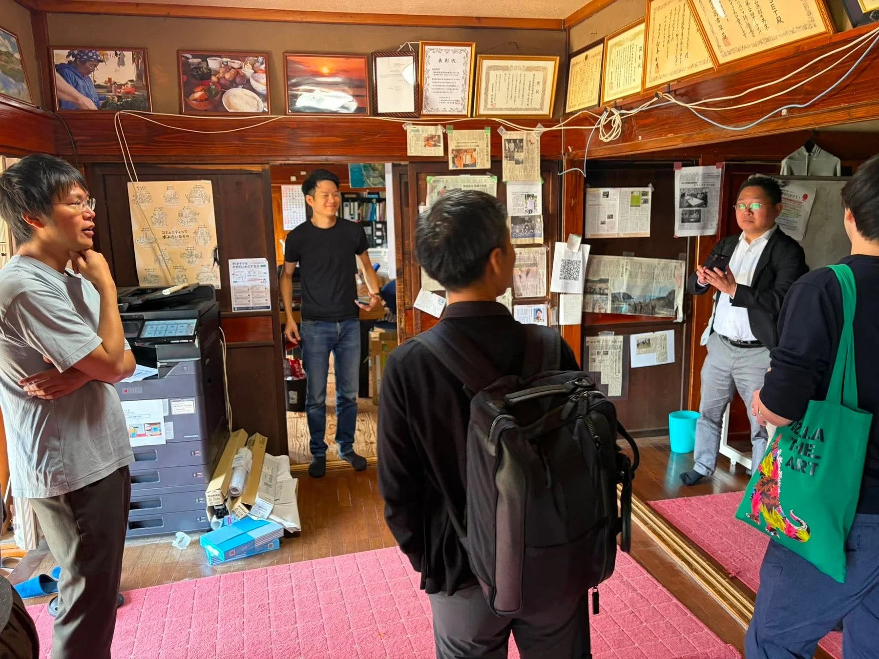 壁に多くの書類や写真が飾られた和風の部屋で、複数の男性が集まって打ち合わせをしている様子