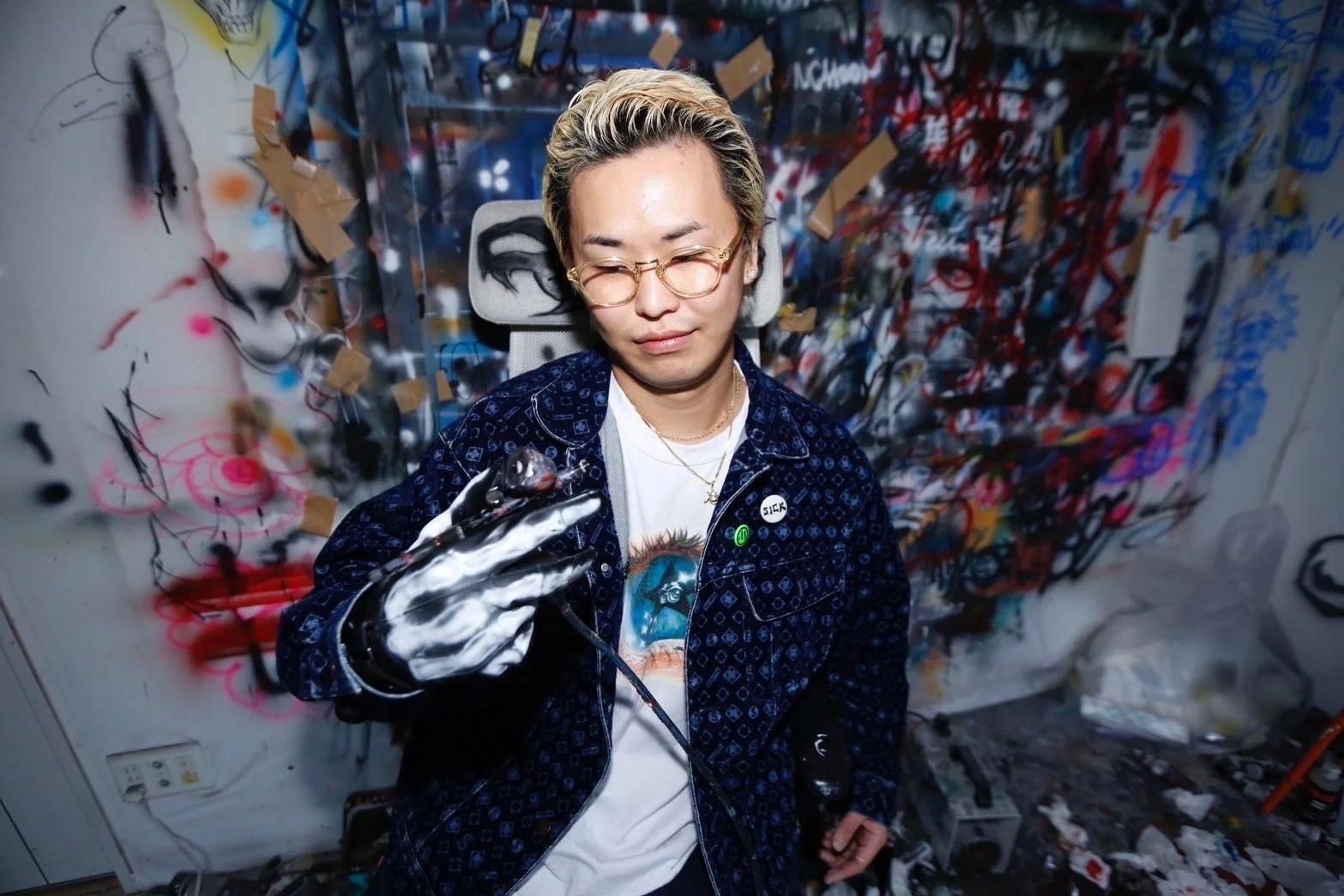 金髪と眼鏡の男性がエアブラシのような道具を持ち、グラフィティで埋め尽くされたアートスタジオに立っている