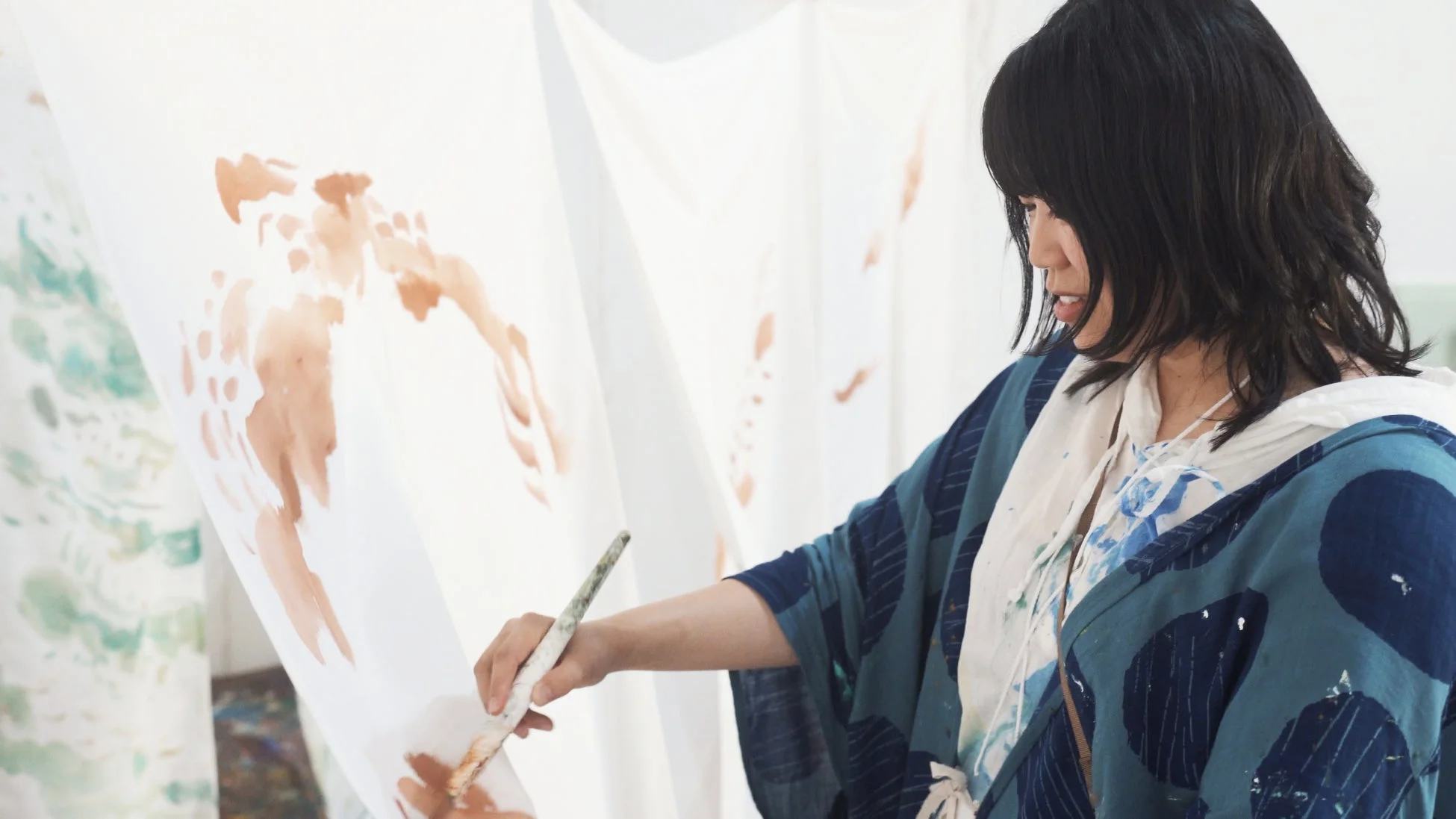 女性が大きな白い布に絵の具で模様を描いている様子を捉えた画像です。彼女は青い模様の入った上着を着用し、真剣な表情で筆を動かしています。