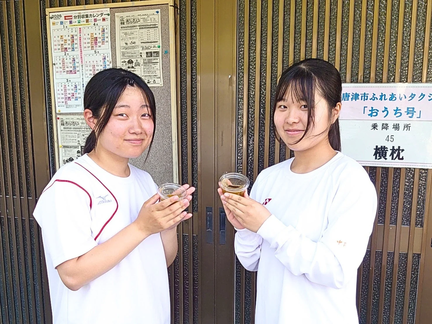 白いスポーツウェアを着た2人の若い女性が、それぞれ小さなカップを持って笑顔で立っています。