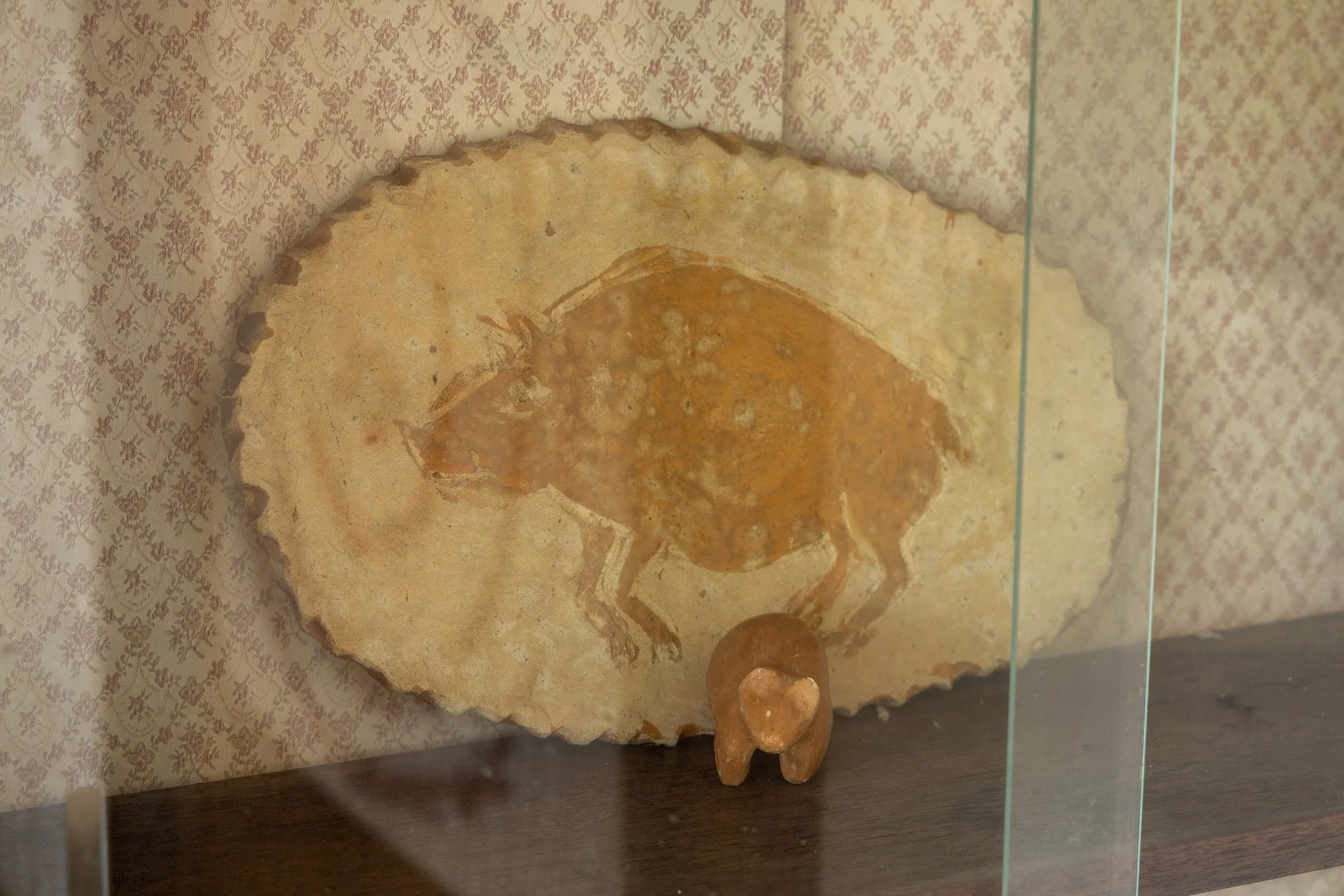 ガラスケースの中に、イノシシの絵が描かれた素朴なレリーフと小さな動物の彫刻が展示されています。背景の模様入り壁紙がレトロな雰囲気を醸し出しています。