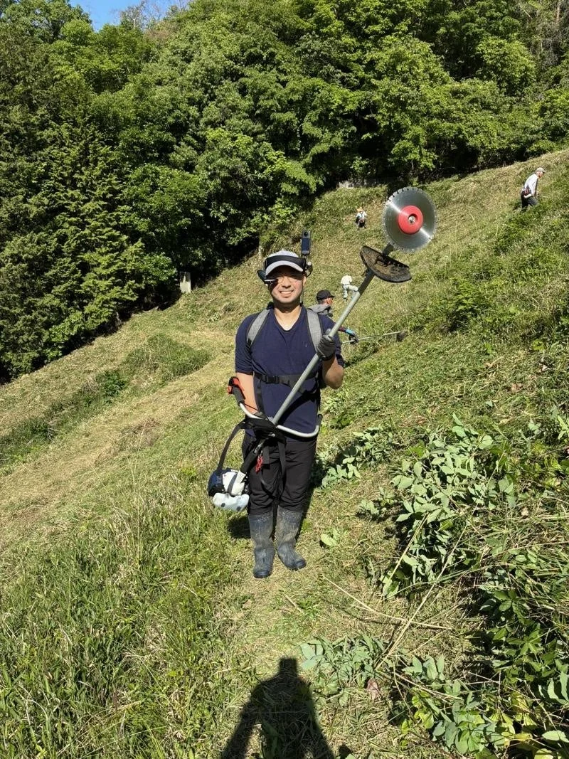 晴れた日に、男性が草刈り機を肩にかけて急な斜面で草刈り作業をしている様子です。男性は笑顔でカメラを見ており、背景には他の作業者や豊かな緑が見えます。