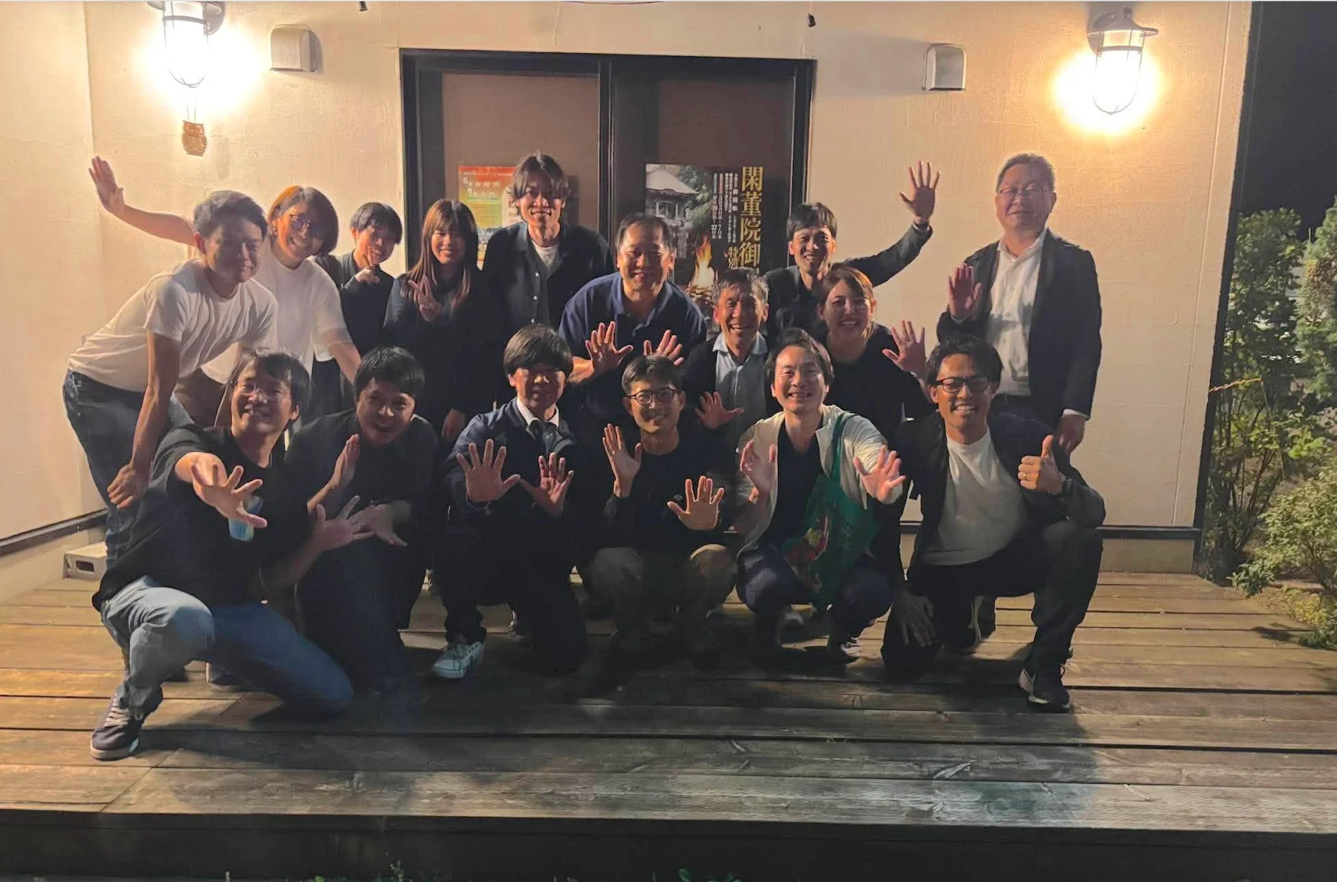 夜の屋外で、多くの人々が木製のデッキに集まり、笑顔でカメラに向かってポーズを取っている集合写真
