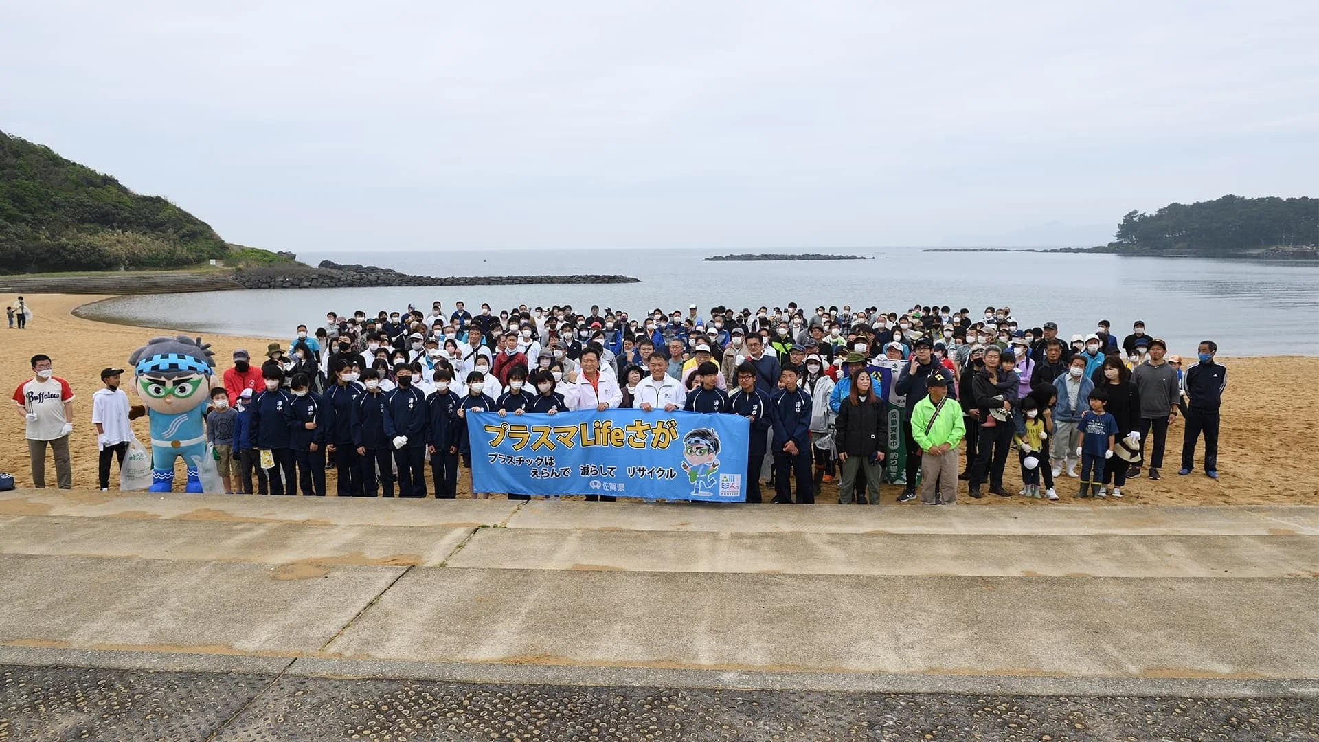 佐賀県の海岸で、マスコットキャラクターと共に多くの人々が集まり、プラスチックごみの削減とリサイクルを呼びかける環境活動を行っている様子。