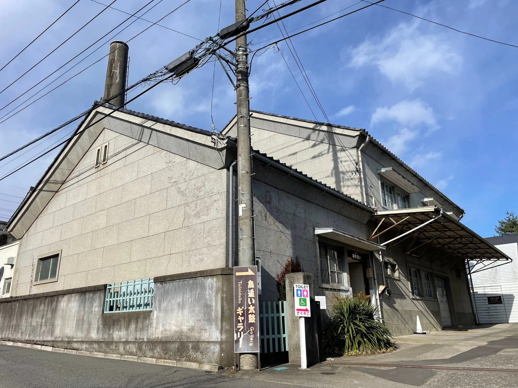 電柱と電線が目立つ、石造りのような外壁を持つ日本の建物。鳥追い太鼓ギャラリーとトイレの案内表示が見える。青空の下、静かな日本の街角の雰囲気を写している。