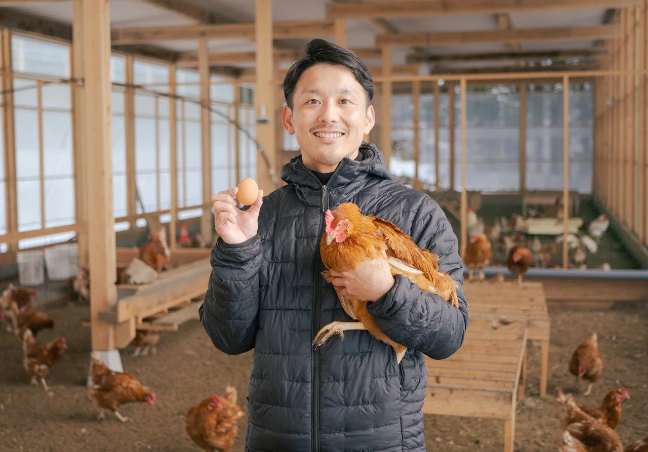 養鶏場で笑顔の男性が鶏を抱え、もう一方の手には新鮮な卵を持っています。