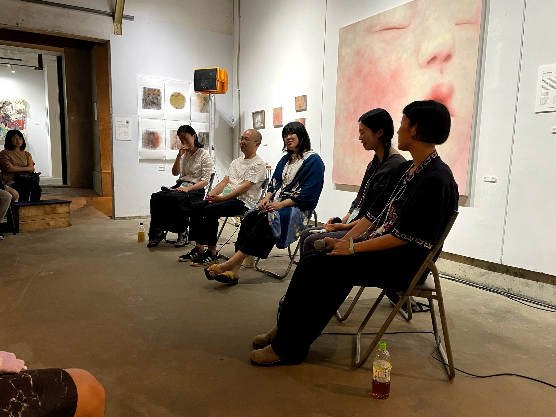 アートギャラリーで開かれているパネルディスカッションかアーティストトークの様子です。数人の人物が椅子に座って話しており、壁には様々な現代アート作品が展示されています。特に右側には大きなピンク色の絵画が見えます。