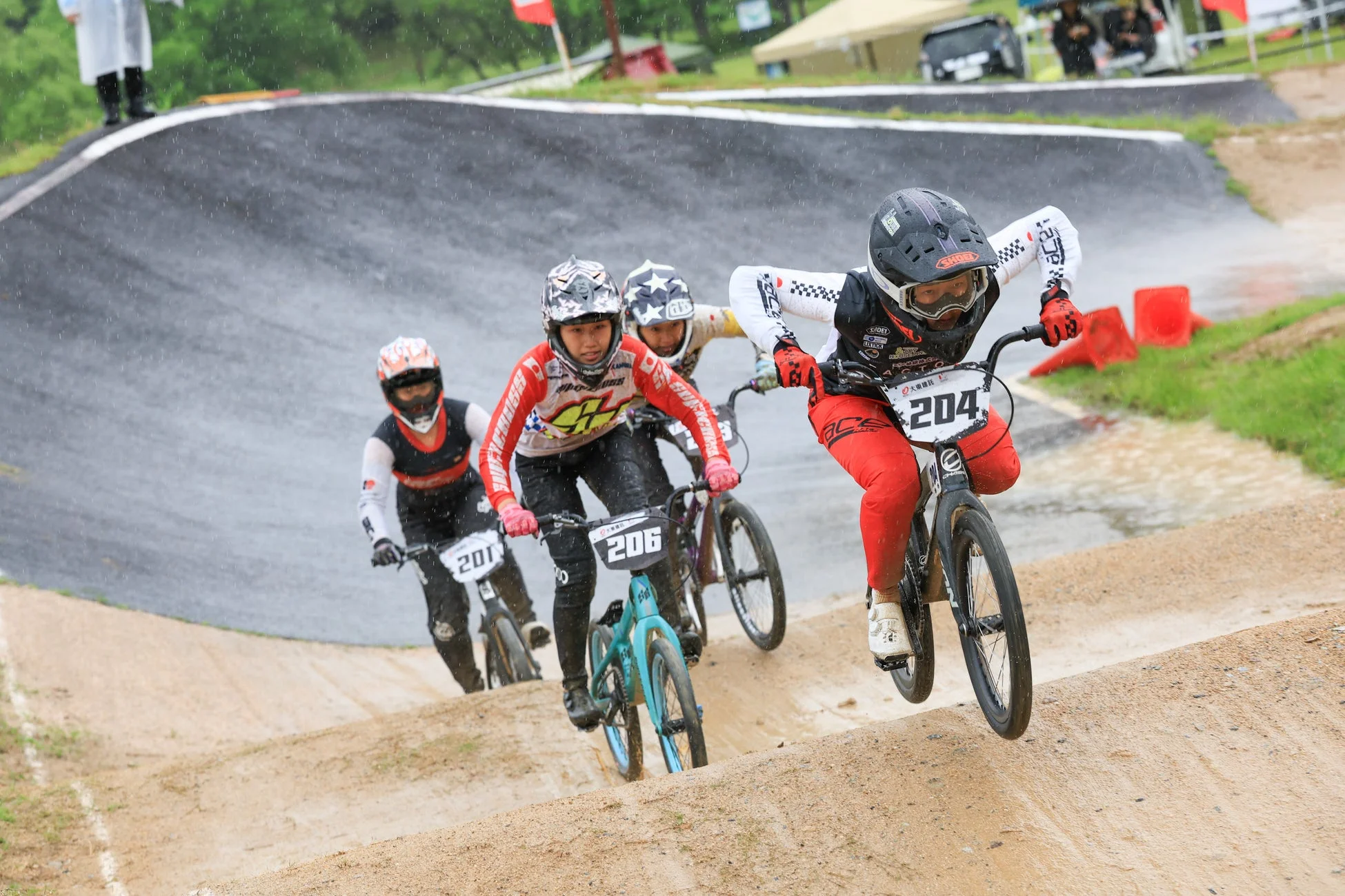 雨が降るダートトラックでBMXレースが行われ、複数の選手が激しく競い合う
