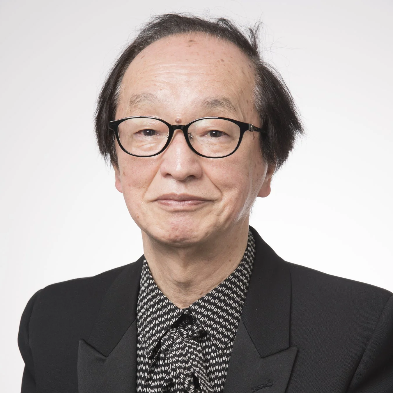 黒縁メガネをかけたアジア系の男性が、黒いスーツと柄物のシャツを着て、穏やかな表情