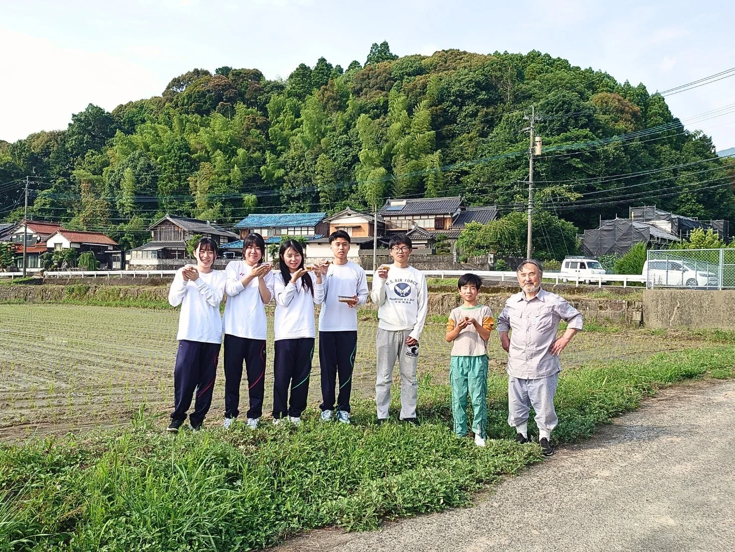 田んぼと日本の伝統的な家屋を背景に、学生服を着た若者たち、子供、大人の混合グループが並んで立っている。