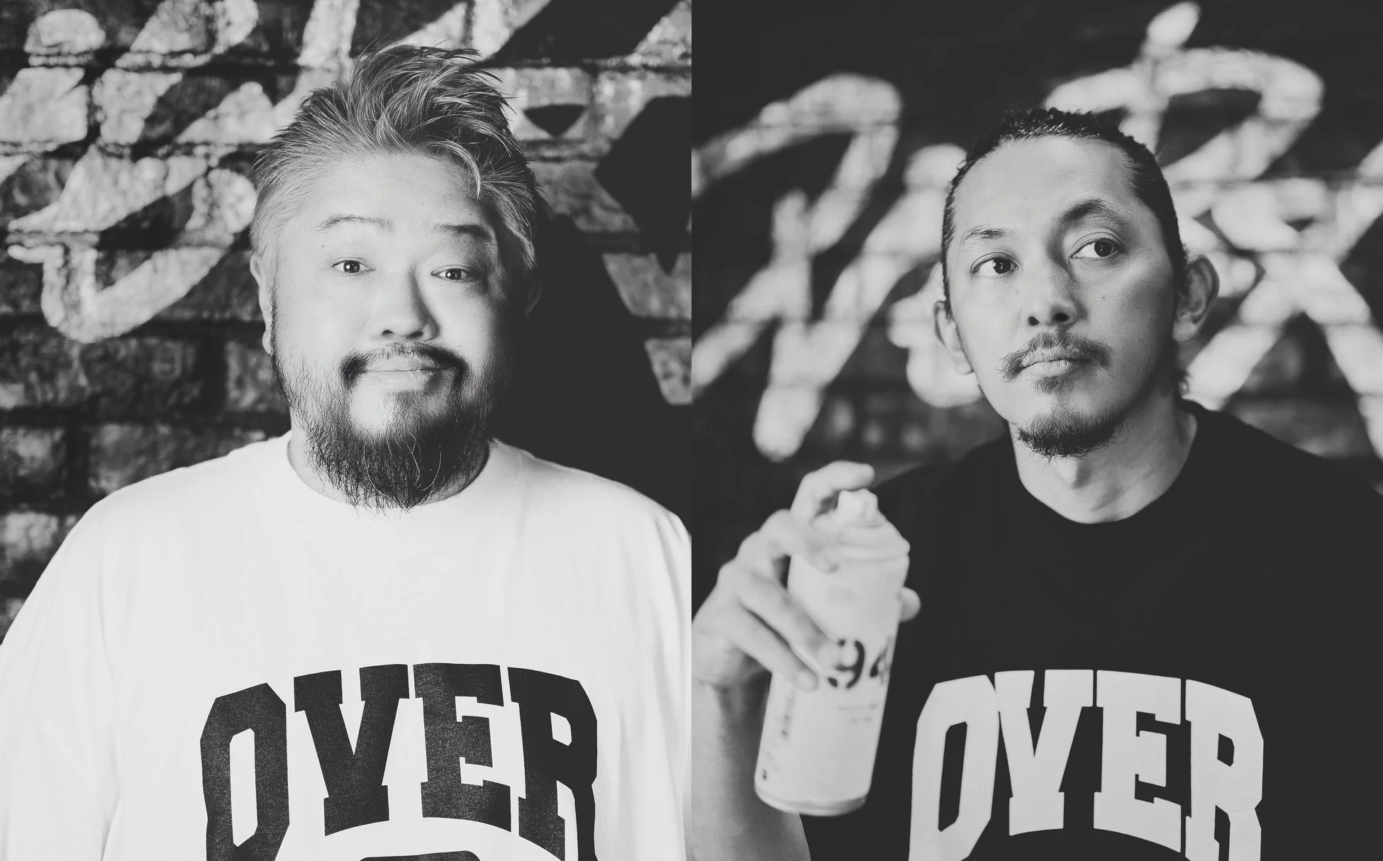 グラフィティが描かれたレンガの壁を背景に、2人の男性が並んで写る白黒写真