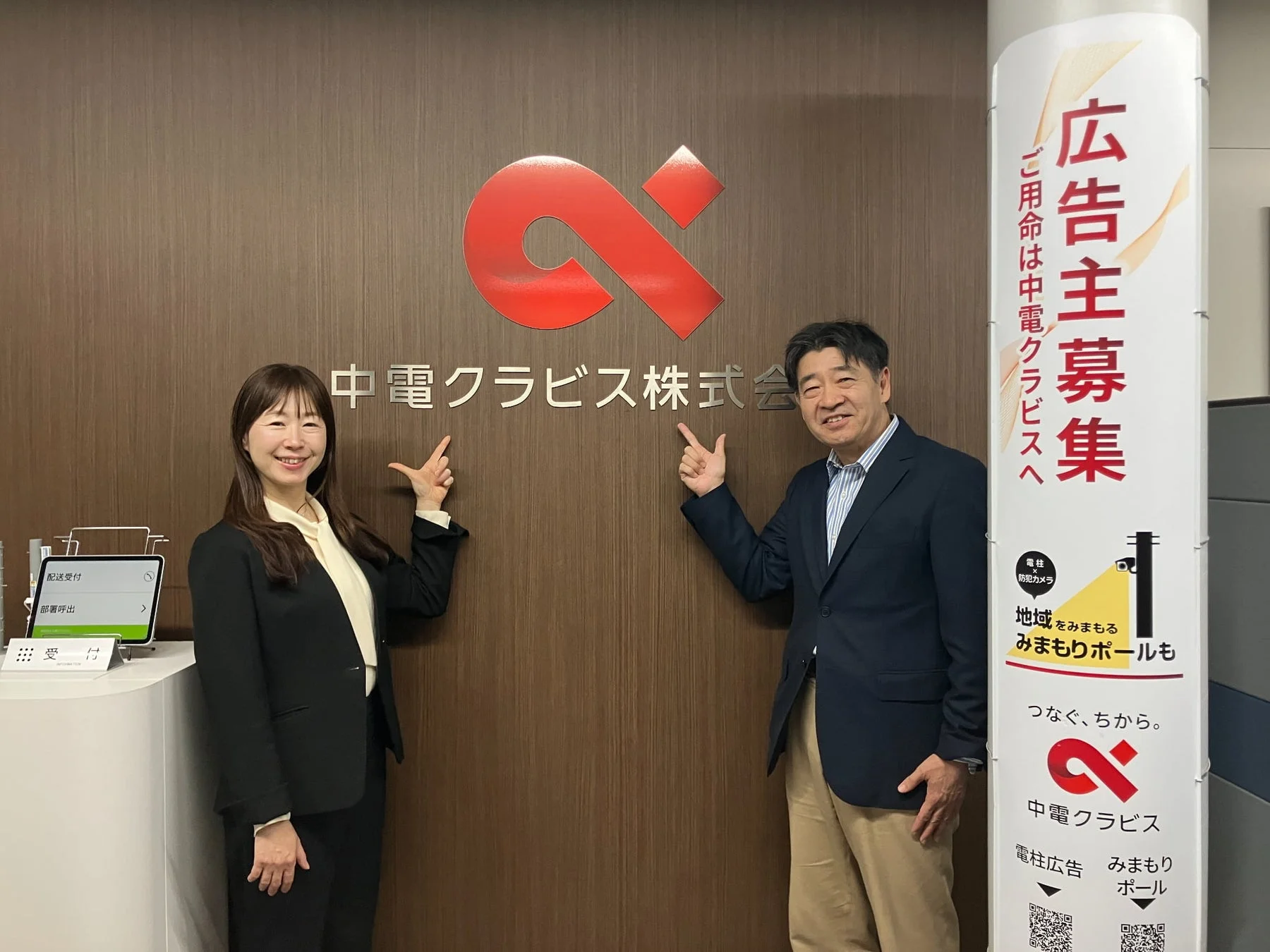 中電クラビス株式会社のオフィスで、笑顔の男女社員が会社名とロゴを指差しています。隣には電柱広告とみまもりポールのサービスを宣伝するバナーがあり、受付のタブレットも写っています。