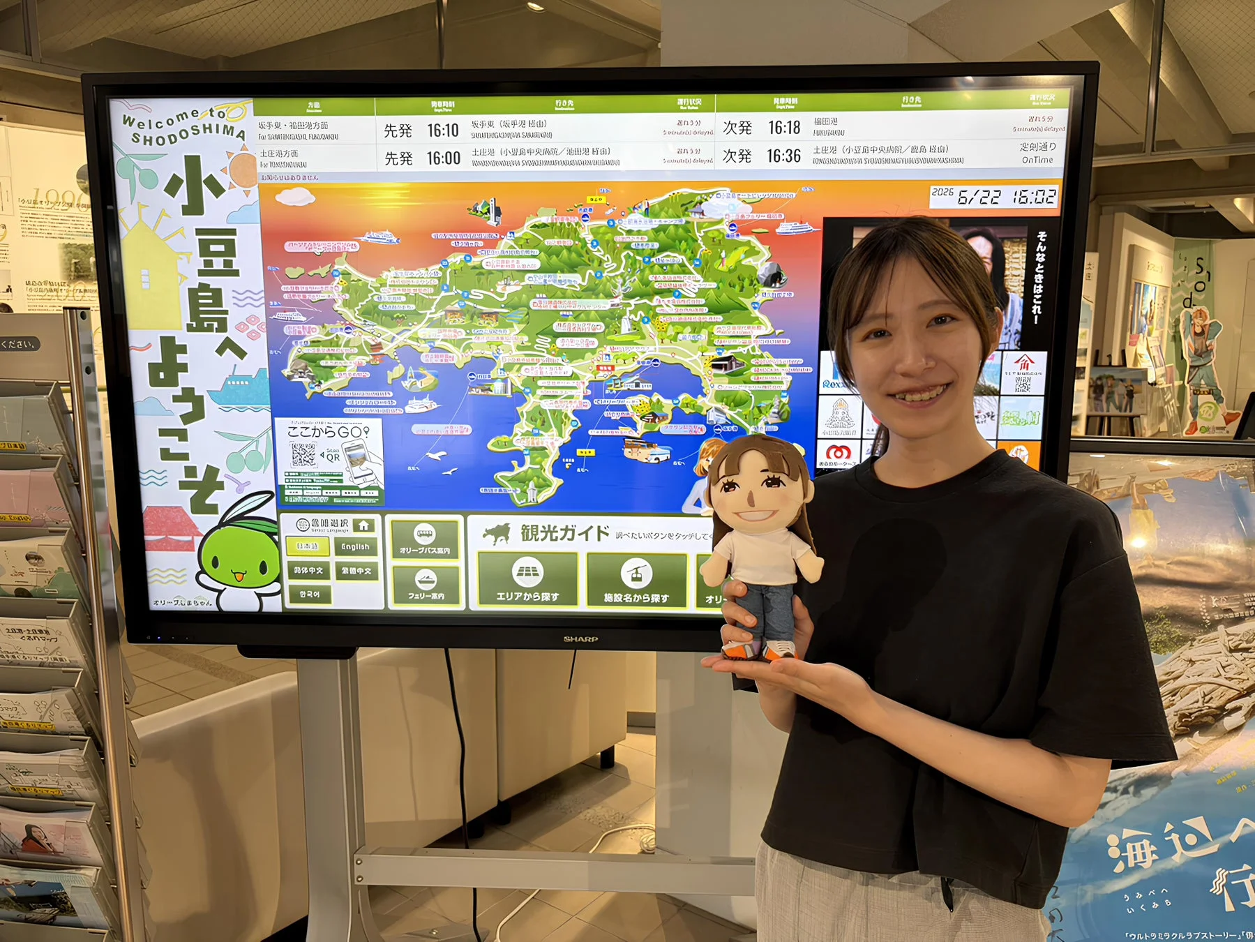 小豆島の観光案内デジタルサイネージの前で、女性が笑顔で人形を抱えています。