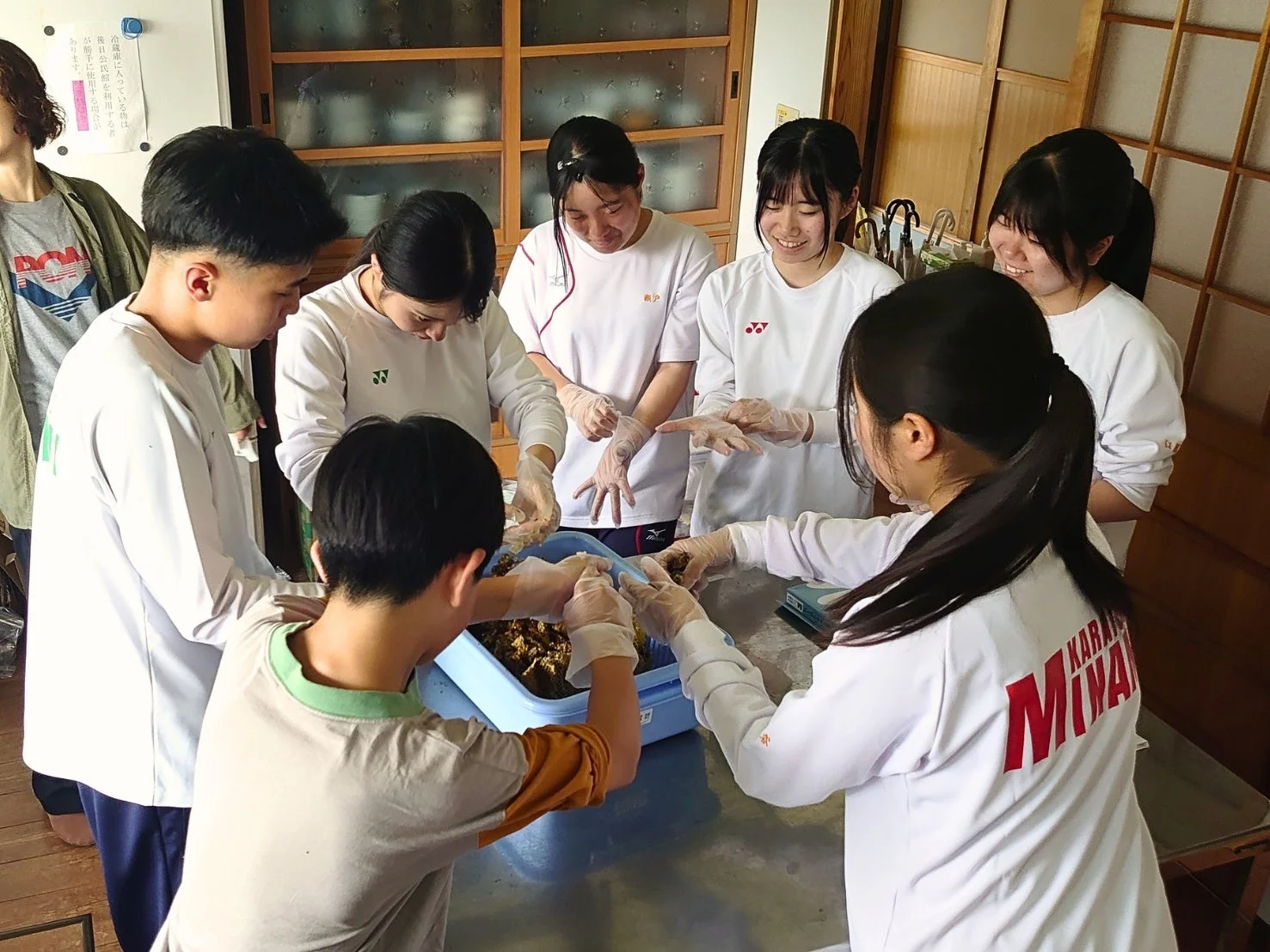 手袋を着用した複数の若者が集まり、青い容器に入った食材を協力して準備している様子。