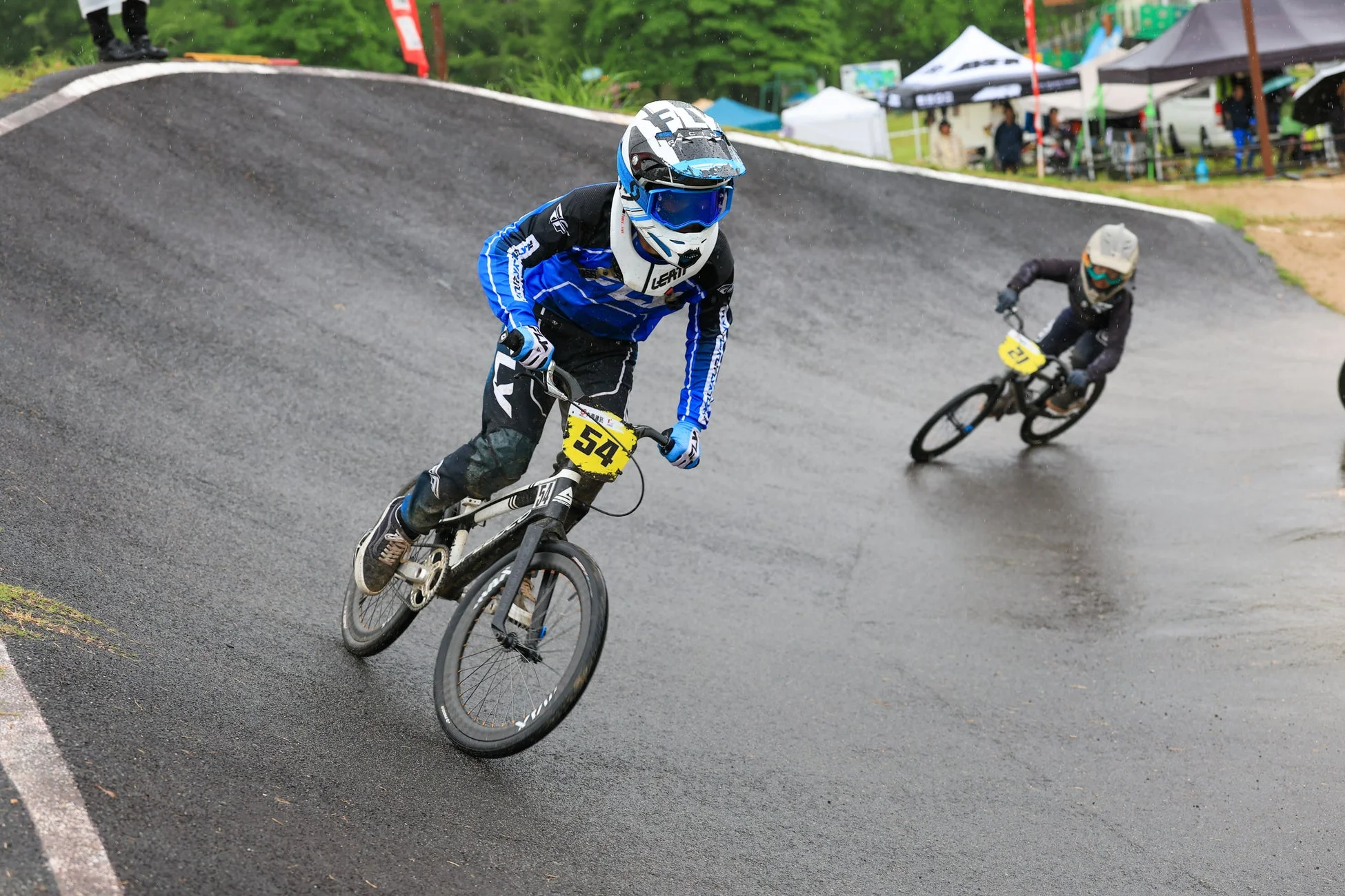 雨が降る屋外のBMXトラックで、2人のライダーが自転車レースをしている様子