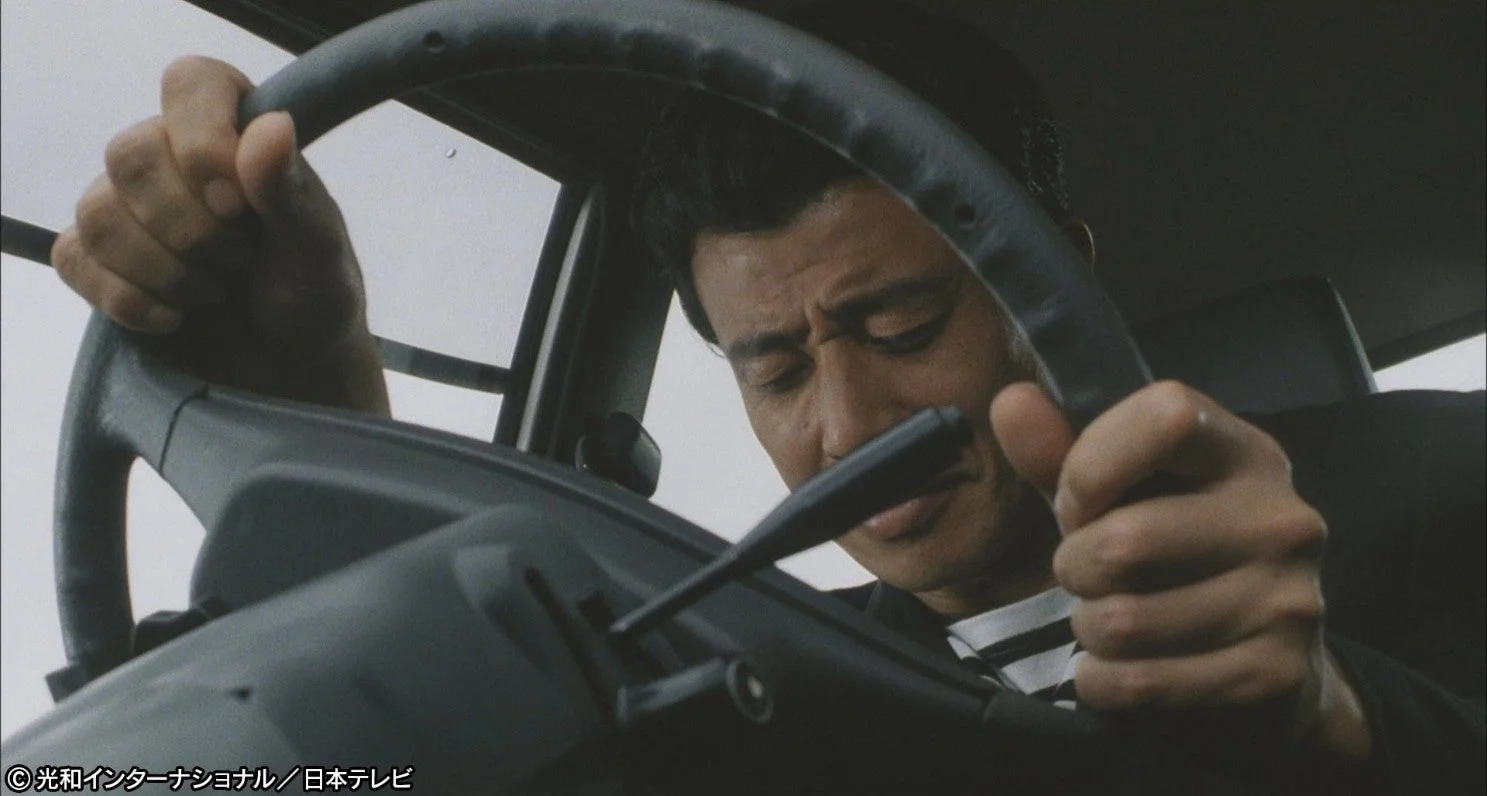 自動車の運転席で、男性がハンドルを強く握りしめ、眉間にしわを寄せて集中しているような、あるいは緊迫した表情を見せている様子です。