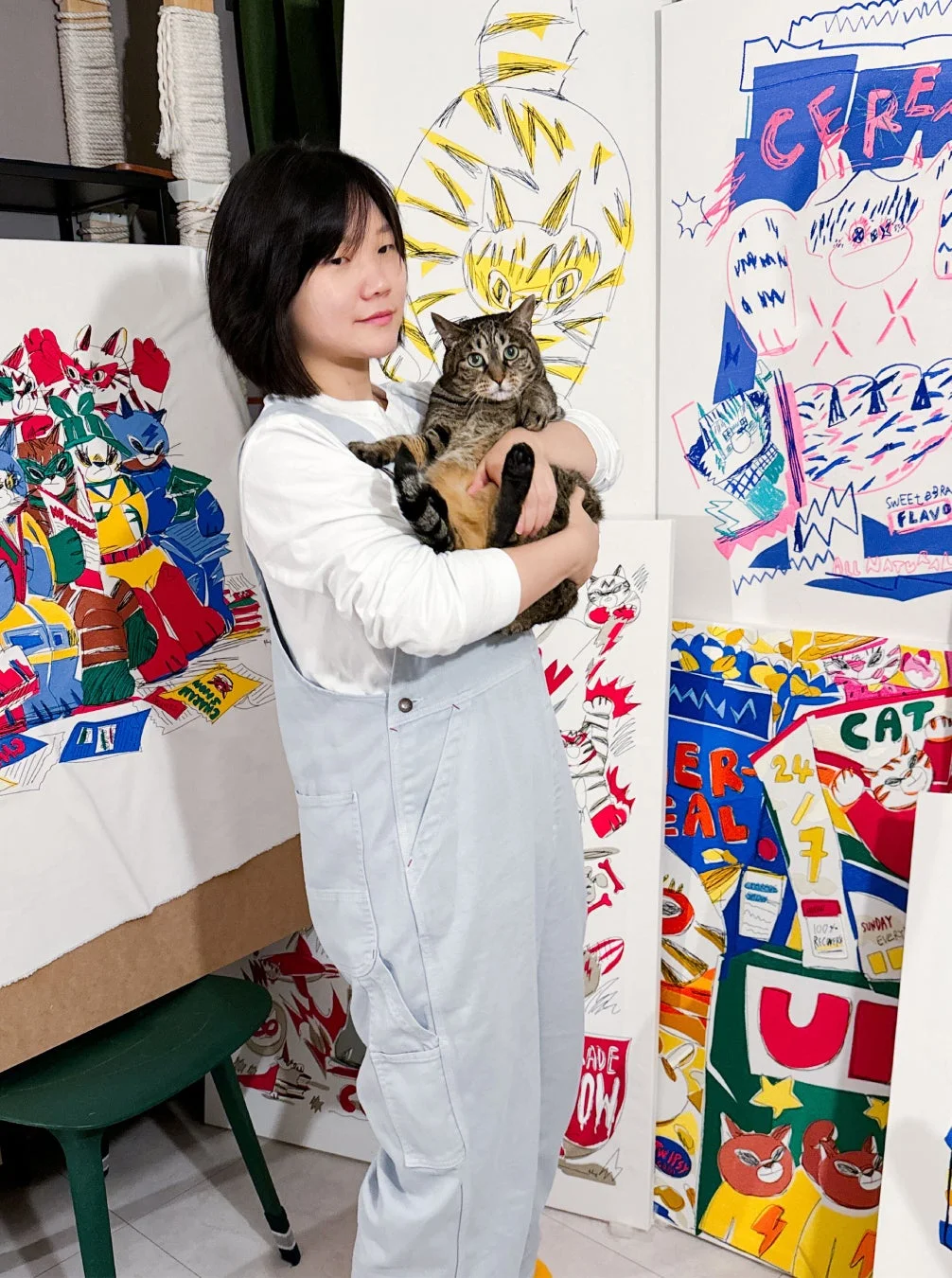 オーバーオールを着た若い女性がキジトラ猫を抱き、周りには猫や抽象的なデザインが描かれたカラフルなアート作品が並ぶアトリエのような空間