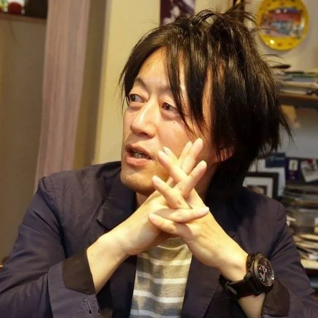 乱れた黒髪の男性が、室内で腕を組み、何かを真剣に話しているか考えているような表情