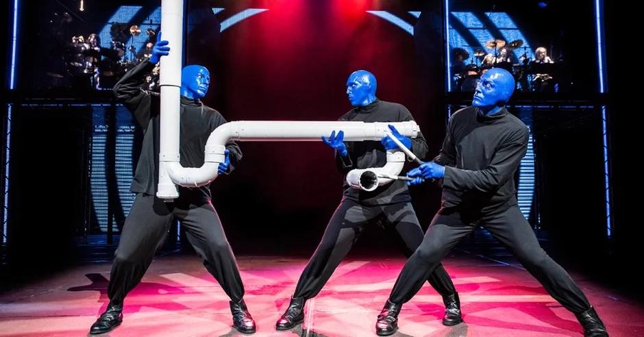 青い顔と黒い衣装の3人のパフォーマーが、白いパイプを楽器のように持って演奏しているステージ