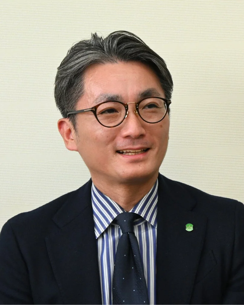 眼鏡をかけたアジア系の男性が、紺色のスーツとストライプのシャツ、ネクタイを着用し、穏やかな笑顔で写っている。